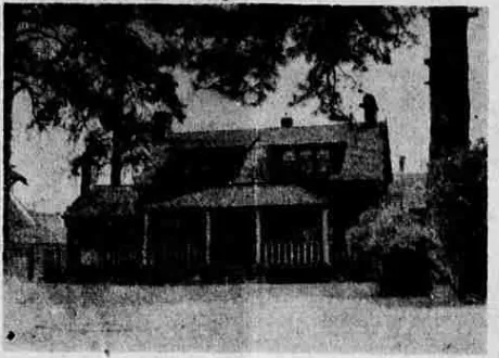 A newspaper photograph of a low house with a shingled roof and a dormer, a deep porch on turned posts and a picket fence, under large trees.