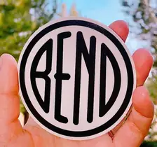 A hand holding a round sticker: a pale disc with a heavy black ring and the word BEND across it in narrow black letters.