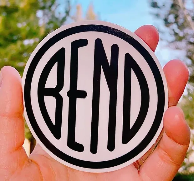 A hand holding a round sticker: a pale disc with a heavy black ring and the word BEND across it in narrow black letters.