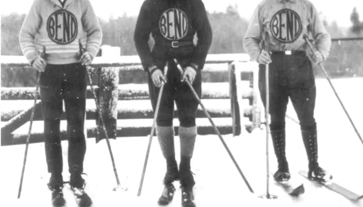 Three people in knitted sweaters and breeches stand on skis in snow before a rail fence, each with a circular BEND emblem on the chest.