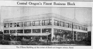 A newspaper photograph of the two-story O’Kane Building at a street corner, with storefronts below its upper windows.