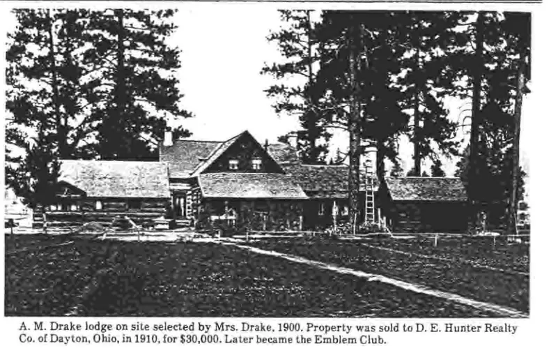A log lodge with a steep roof, chimneys and a porch, beneath tall trees.