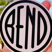 A hand holding a round sticker: a pale disc with a heavy black ring and the word BEND across it in narrow black letters.